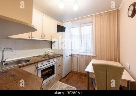 cuisine confortable avec des armoires beiges, des plans de travail sombres et une zone de repas près d'une fenêtre. Les appareils comprennent une cuisinière, un four et un réfrigérateur Banque D'Images