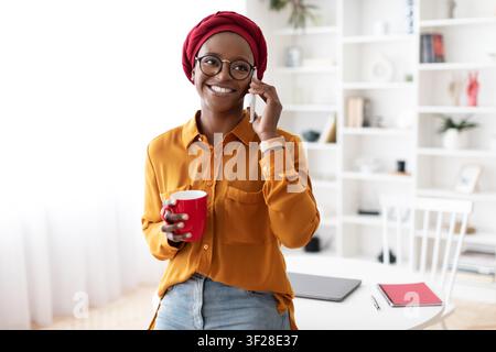 Femme afro-américaine heureuse téléphonant tout en tenant une tasse dans un bureau moderne à domicile Banque D'Images