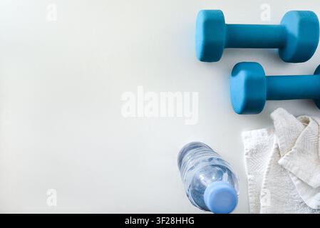 Scène de fitness minimaliste avec des haltères bleus, une serviette blanche et une bouteille d'eau sur un fond lumineux. Composition épurée offrant un espace de copie généreux f Banque D'Images