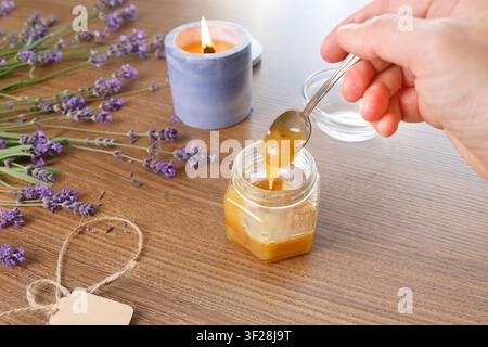 Main humaine tenant une cuillère versant du miel biologique d’un bocal sur une table en bois en arrière-plan. Bougie à la lavande brûlante, bouquet de fleurs violettes, vue de dessus. Confortable Banque D'Images