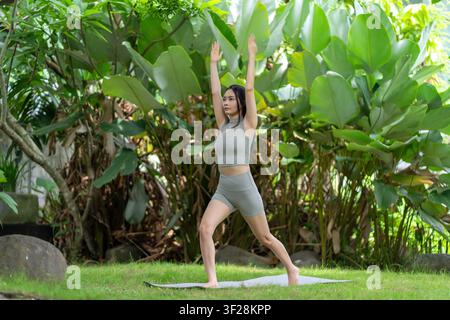 Une Indonésienne réalise une pose de yoga d'équilibre sur un tapis dans un jardin luxuriant en extérieur, mettant en valeur le calme, l'exercice et le bien-être naturel. Banque D'Images