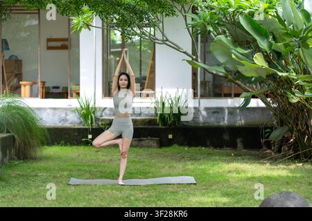 Une Indonésienne réalise une pose de yoga d'équilibre sur un tapis dans un jardin luxuriant en extérieur, mettant en valeur le calme, l'exercice et le bien-être naturel. Banque D'Images
