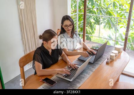Deux femmes d'affaires asiatiques collaborant sur des ordinateurs portables dans un espace de travail moderne et lumineux avec lumière naturelle. Banque D'Images