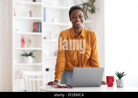 Femme noire souriante debout à la table du bureau à domicile avec ordinateur portable, concept commercial Banque D'Images
