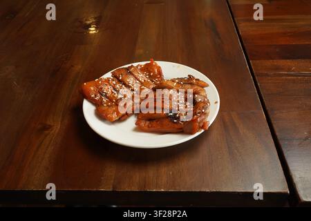 Tranches de poulet teriyaki grillées garnies de graines de sésame servies dans une assiette blanche sur une table en bois de restaurant. Banque D'Images