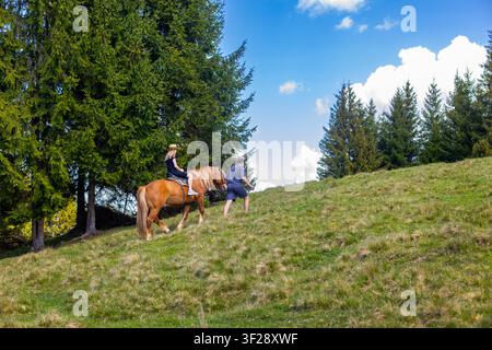 Femme montant un cheval sur une colline herbeuse. Une femme monte un cheval bai tandis qu'un homme marche devant elle sur une colline herbeuse ensoleillée bordée de grands Banque D'Images