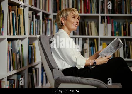 Femme caucasienne d'âge moyen lisant un document imprimé dans une bibliothèque à domicile, examinant des informations et souriant. Utile pour l'éducation, l'édition, la recherche, les commentaires d'experts Banque D'Images