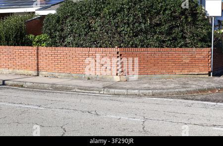 Clôture en briques au coin de la rue. Plante à feuilles persistantes derrière, trottoir et rue devant. Arrière-plan urbain pour espace de copie. Banque D'Images