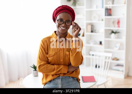Femme afro-américaine souriante avec des lunettes posant avec assurance dans un bureau moderne à domicile Banque D'Images