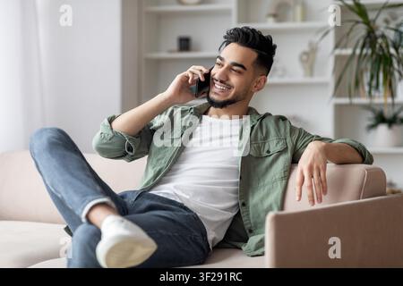 Jeune homme heureux téléphonant tout en se détendant sur un canapé dans un salon moderne Banque D'Images