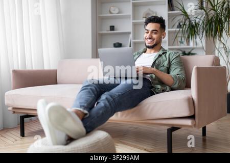 Jeune homme hispanique souriant utilisant un ordinateur portable tout en se détendant sur un canapé à la maison, concept de travail à distance Banque D'Images