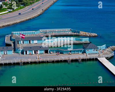 Vue aérienne de Skovshoved Havbad avec des gens bronzant sur la terrasse en bois et le drapeau danois flottant, entouré par une mer bleue claire et une route côtière Banque D'Images