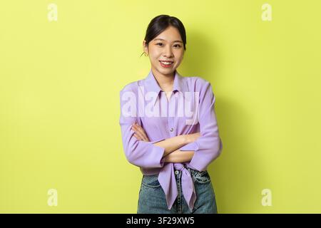 Femme asiatique souriante aux bras croisés posant avec assurance, fond vert, concept commercial Banque D'Images