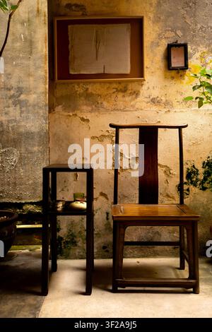 Prise verticale d'une chaise en bois chinoise classique et d'une table d'appoint contre un mur rustique et patiné, décoré d'œuvres d'art encadrées dans une cour Banque D'Images