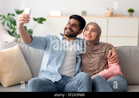 Jeune couple heureux prenant un selfie ensemble à la maison, souriant et se détendant sur le canapé Banque D'Images