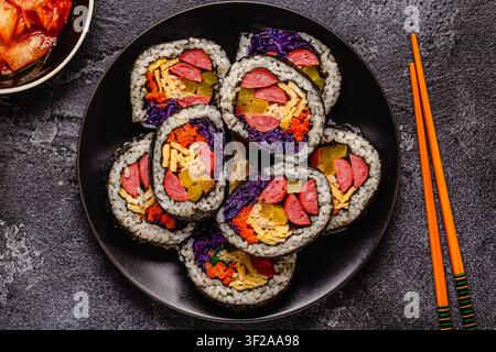 Une assiette noire remplie de kimbap (ou gimbap) aux garnitures colorées. Les ingrédients comprennent des saucisses, des cornichons, des œufs et des légumes. Banque D'Images