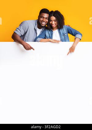 Homme et femme afro-américains souriants pointant vers une pancarte vierge, fond jaune, concept publicitaire Banque D'Images