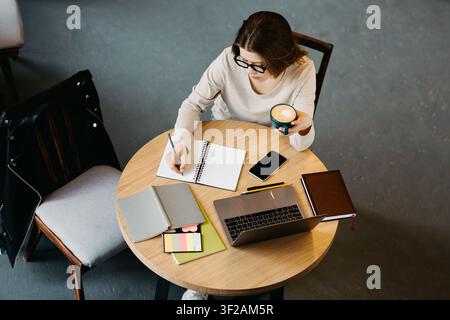 Jeune femme travaillant dans un café avec un ordinateur portable et un bloc-notes, concentrée sur ses notes, concept commercial Banque D'Images