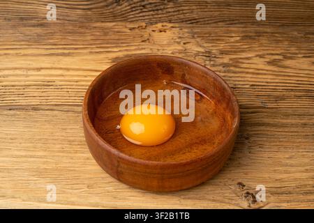 Un œuf cassé dans un bol en bois, jaune d’œuf et blanc crus, œuf de poule biologique fraîchement cassé sur un fond rustique en bois Banque D'Images