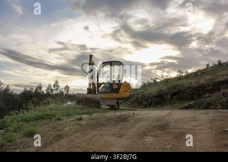 Une petite pelleteuse sur chenilles jaune est à l'arrêt sur un chemin de terre sur une colline entourée d'arbres et de végétation verte sous un ciel nuageux dramatique. Banque D'Images