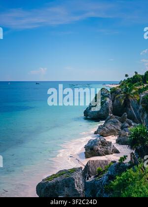 Ciel bleu vif rencontre une mer turquoise. Des falaises abruptes encadrent une plage douce baignée de soleil. Une végétation luxuriante grimpe sur des affleurements rocheux. Des bateaux lointains parsèment les eaux calmes. Paix Banque D'Images