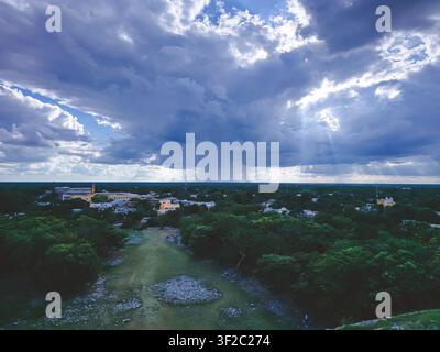 Des nuages dramatiques planent au-dessus d'une ville tranquille. Les rayons du soleil traversent le ciel, projetant des rayons dorés. Des arbres verts et luxuriants recouvrent le paysage en contrebas. Un sentier de terre serpente Banque D'Images