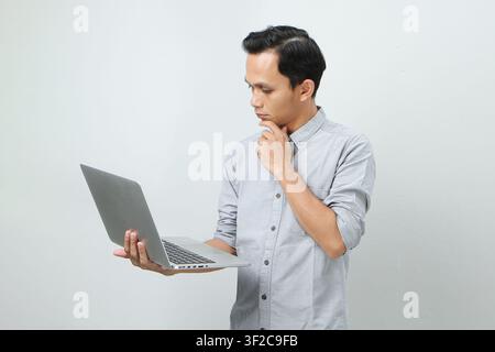 Jeune homme indonésien asiatique pensif tenant un ordinateur portable avec un visage sérieux, réfléchissant à une question, pensif face à une idée déroutante sur un fond isolé Banque D'Images