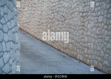 Vue rapprochée d'un mur de pierre et d'un chemin en béton adjacent, mettant en valeur une surface texturée extérieure avec des pierres naturelles disposées selon des motifs irréguliers Banque D'Images