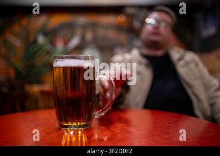 Verre de bière légère sur une table avec un homme flou tendant la main vers la boisson dans un pub. Boisson alcoolisée rafraîchissante, atmosphère de bar, vie nocturne, loisir Banque D'Images