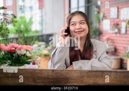 Un plan moyen d'une jeune femme d'affaires thaïe du Sud-Est de l'Isan portant une veste de costume grise, tenant un téléphone portable à son oreille et souriant tout en énumérant Banque D'Images