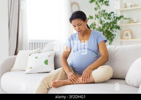 Femme noire enceinte assise sur un canapé et massant sa cheville, montrant une douleur à la jambe Banque D'Images