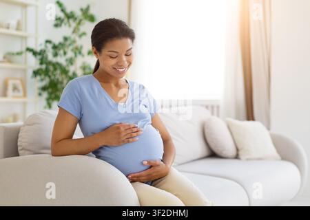Femme noire enceinte assise sur un canapé, berçant son ventre avec les deux mains Banque D'Images