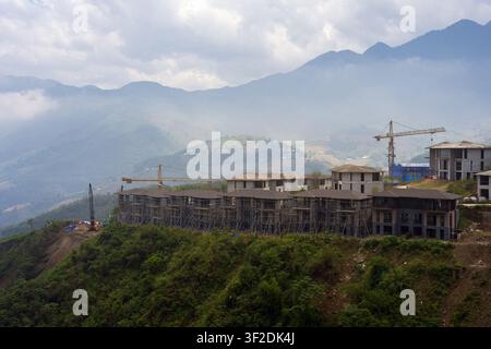 Des bâtiments à plusieurs étages en construction surplombent des pentes montagneuses accidentées et des nuages dérivants près de Sa Pa, Lao Cai, Vietnam. Banque D'Images