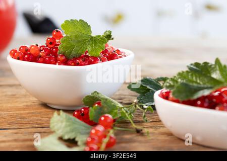 Groseilles rouges vives débordant d'un bol en céramique blanche avec une feuille verte brillante, brins épars et surface en bois rustique, lumière naturelle douce, nature morte de fruits d'été frais et éclatants avec des couleurs riches et Banque D'Images