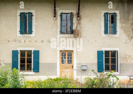 Façade de maison rustique érodée avec volets bleu-vert et une porte en bois, fenêtres symétriques, plâtre écaillé et plantes grimpantes, entourée de verdure Banque D'Images