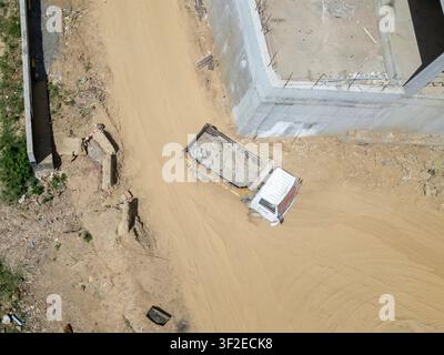 Vue aérienne d'un chantier de construction montrant un camion chargé de débris de béton. Le site est entouré d'un chemin de terre et de structures partielles, indiquant Banque D'Images