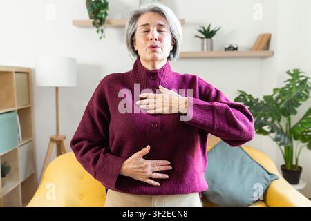 Sénior femme pratique la respiration consciente à la maison, main sur la poitrine et le ventre, se concentrant sur une routine de bien-être calme dans un salon lumineux avec des plantes et s Banque D'Images
