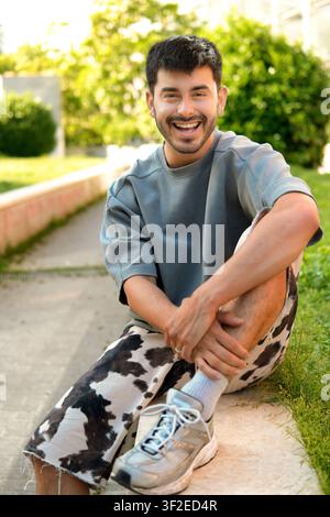 Homme hispanique souriant et à la mode assis sur un trottoir urbain ensoleillé, portant une tenue de streetwear et des baskets. Portrait de style de vie décontracté avec de la verdure an Banque D'Images