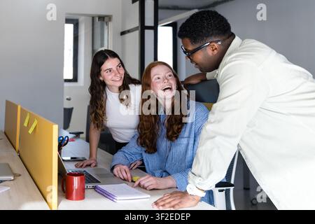 Des collègues divers collaborent à un bureau dans un bureau moderne, souriant et riant tout en discutant d'un projet sur un ordinateur portable avec des notes, une tasse de café et br Banque D'Images