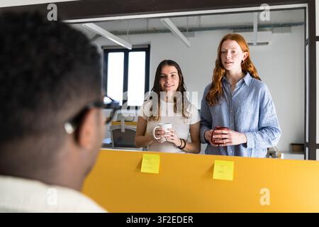 Deux collègues tiennent des tasses de café et discutent par-dessus un bureau dans un bureau moderne et lumineux, avec des post-it sur une cloison et un collègue au premier plan tenant une liste Banque D'Images