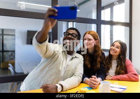 Trois collègues divers prennent un selfie à une table jaune dans un bureau moderne aux murs de verre, souriants après une réunion. Moment de travail d'équipe décontracté avec smartphone Banque D'Images