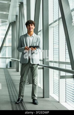Portrait en pleine longueur d'un jeune professionnel d'affaires en costume léger tenant une tablette dans un couloir vitré moderne et lumineux, suggérant la technologie, fina Banque D'Images