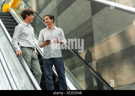 Deux collègues d'affaires descendent un escalier mécanique dans un bâtiment moderne, discutant et souriant. L'un tient un smartphone, suggérant le travail d'équipe, le réseautage, un Banque D'Images