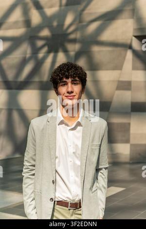 Jeune homme professionnel en costume clair pose à l'extérieur sous un soleil chaud, debout devant un mur géométrique moderne avec des ombres douces, exprimant la confiance Banque D'Images