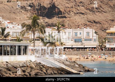 Las Palmas, Îles Canaries, Espagne - 17 juillet 2025 : Vue sur Puerto de Mogan avec des bâtiments blancs, des palmiers et des gens se relaxant sur la plage de sable et Banque D'Images