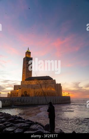 Casablanca, Casablanca-Settat, Maroc - 15 novembre 2025 : Vue de la mosquée Hassan II au coucher du soleil avec un pêcheur debout sur le rivage rocheux au premier plan Banque D'Images