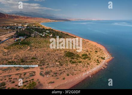 Vue aérienne de la côte de sable roux du lac Issyk-Kul avec un petit village et des montagnes lointaines à Kaji-Say, région d'Issyk-Kul, Kirghizistan. Banque D'Images