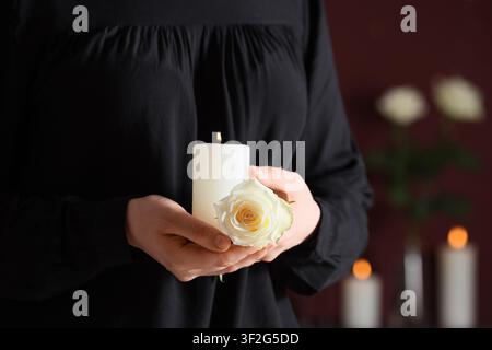 Person en noir tenant une bougie blanche allumée et une délicate rose blanche, deuil et souvenir Banque D'Images