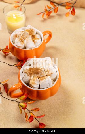 Deux tasses en forme de citrouille remplies de chocolat chaud, garnies de guimauves moelleuses et de cannelle, créant une scène de boisson automnale chaleureuse avec une lumière allumée Banque D'Images