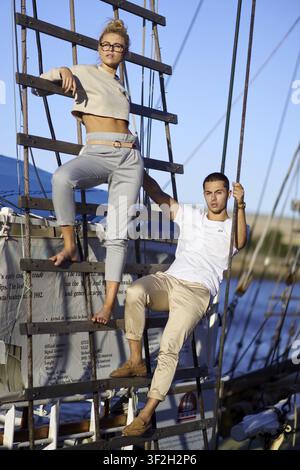 En équilibre suspendu, deux personnes sur une échelle de bateau, l'une debout, l'autre assise, en tenue d'été décontractée, ambiance détendue, couple de marins, capitaine de bateau Banque D'Images